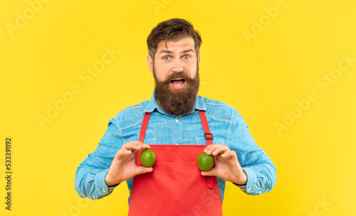Surprised man in red apron holding fresh limes citrus fruits yellow background, greengrocer