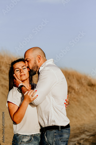 Happy smiling Couple walking at Sand Dunes near the Beach. Young happy Bearded muscular man in White shirt kissing and hugging beautiful woman at sunser on a beach