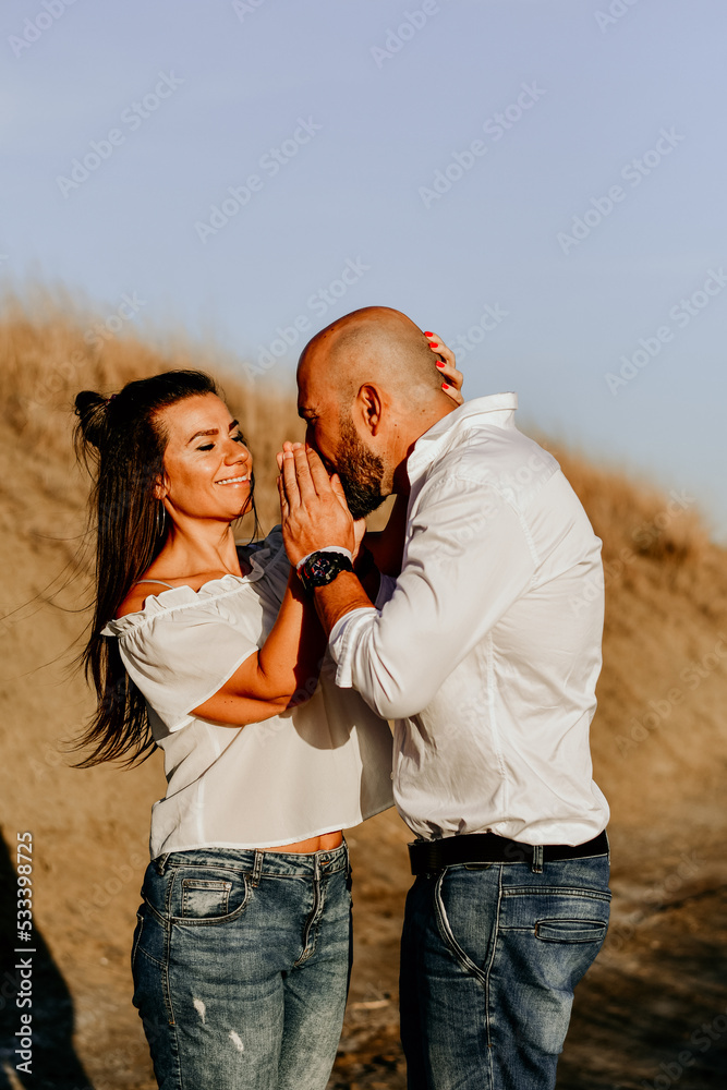 Happy smiling Couple walking at Sand Dunes near the Beach. Young happy Bearded muscular man in White shirt kissing and hugging beautiful woman at sunser on a beach Happy smiling Couple walking at Sand Dunes near the Beach. Young happy Bearded muscular man in White shirt kissing and hugging beautiful woman at sunser on a beach