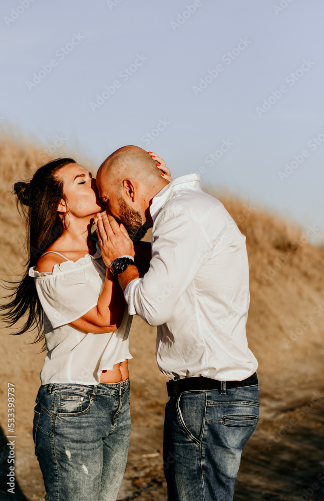 Happy smiling Couple walking at Sand Dunes near the Beach. Young happy Bearded muscular man in White shirt kissing and hugging beautiful woman at sunser on a beach Happy smiling Couple walking at Sand Dunes near the Beach. Young happy Bearded muscular man in White shirt kissing and hugging beautiful woman at sunser on a beach