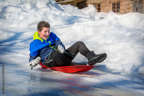 In winter, in a forest, a boy lying on his stomach slides down a hill on a plastic plate. He is joyful.