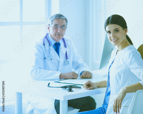 A doctor smiling at the camera with her male colleage in the back of the consulting room in hospital