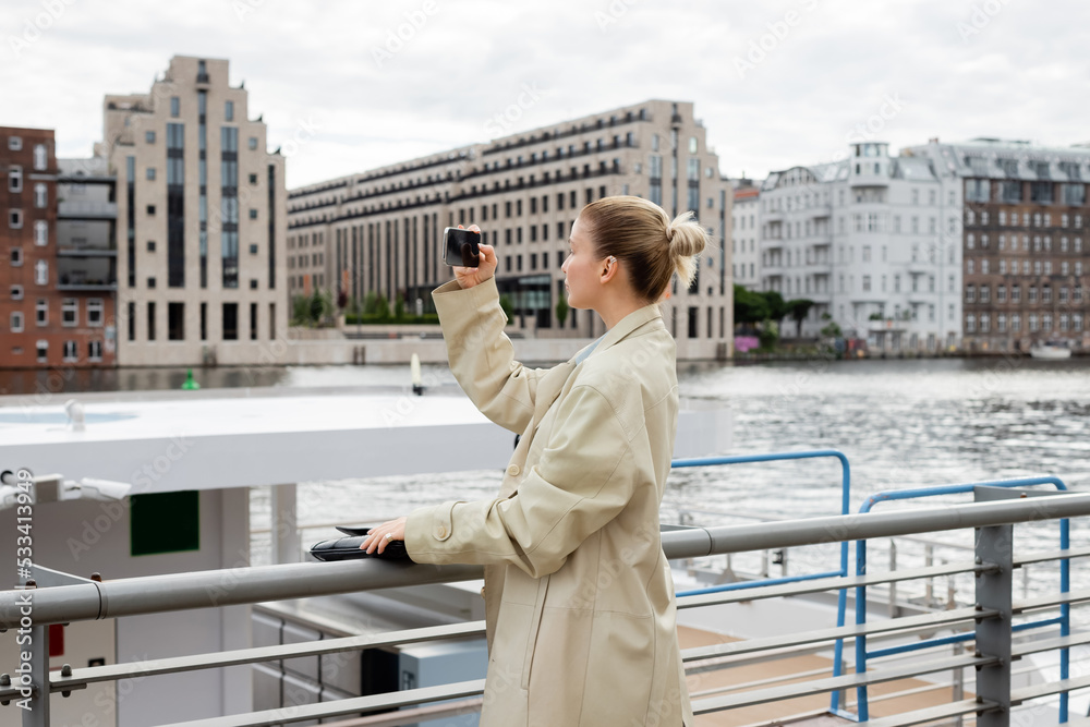 Side view of woman in trench coat taking photo on cellphone near river in Berlin.