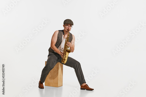 Young musician in retro style outfit playing saxophone isolated over white studio background. Concept of music, hobby, festival.