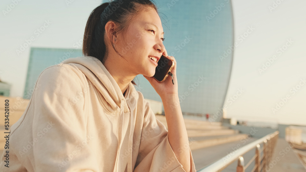 Smiling Asian girl in sportswear talking on the phone while standing on the embankment on modern buildings background in the morning light Smiling Asian girl in sportswear talking on the phone while standing on the embankment on modern buildings background in the morning light
