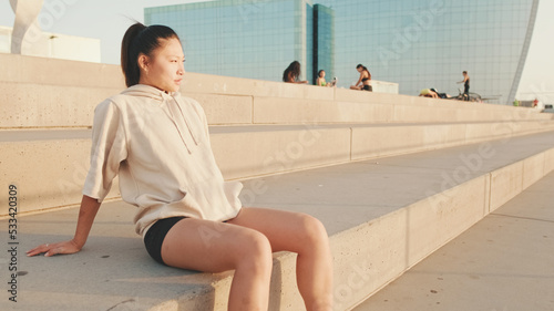 Asian girl in sportswear resting after workout sitting on steps on modern buildings background at sunrise photo