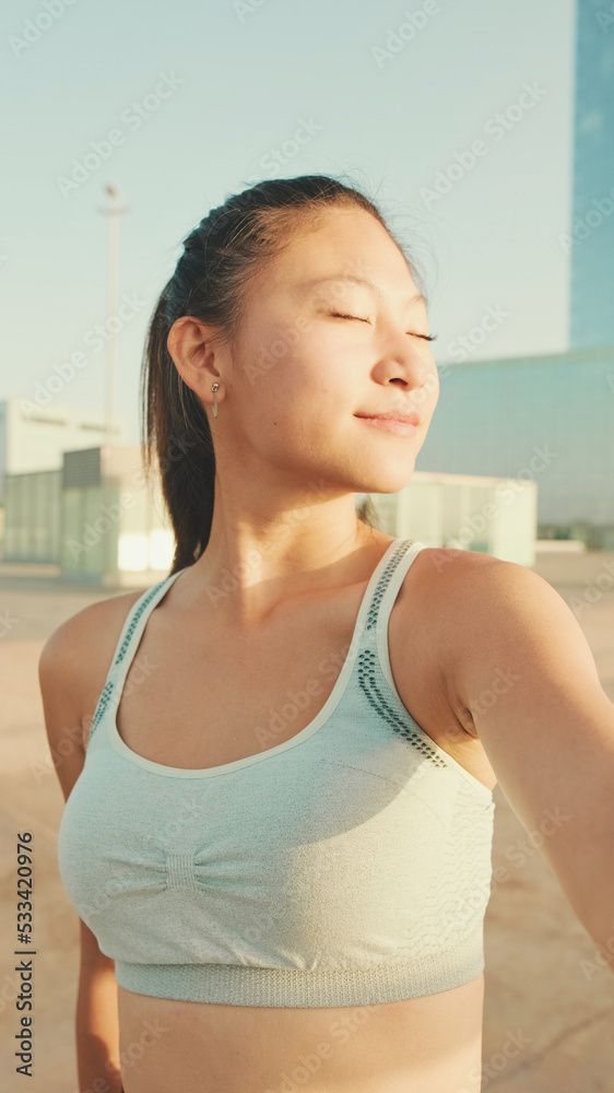 Smiling asian girl in sports top makes selfie while standing on the embankment in the morning light Smiling asian girl in sports top makes selfie while standing on the embankment in the morning light