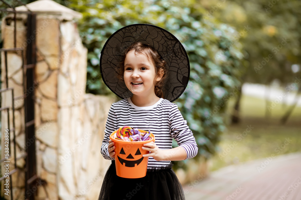 Φωτογραφία Little cute girl in witch costume holding jack-o-lantern pumpkin bucket with candies and sweets