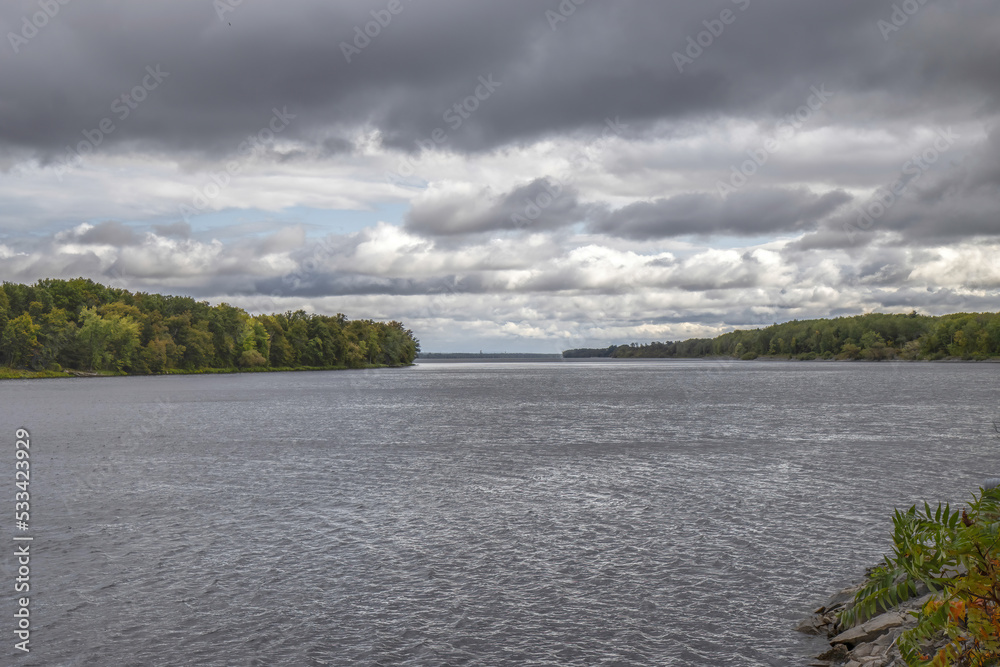 Wide dark cold river moving under a threatening sky with dark clouds, forests on shore, daytime, overcast, nobody