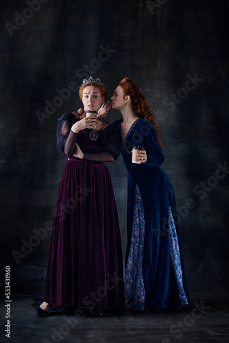 Portrait of two beautiful women in image of queens isolated over dark background. Whispering secrets and gossips