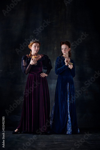 Portrait of two beautiful women in image of queens isolated over dark background. Junk and healthy food