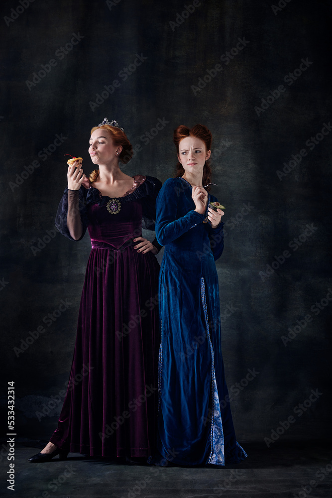Portrait of two beautiful women, royal persons eating cake and avocado isolated over dark background. Junk and healthy food Portrait of two beautiful women, royal persons eating cake and avocado isolated over dark background. Junk and healthy food
