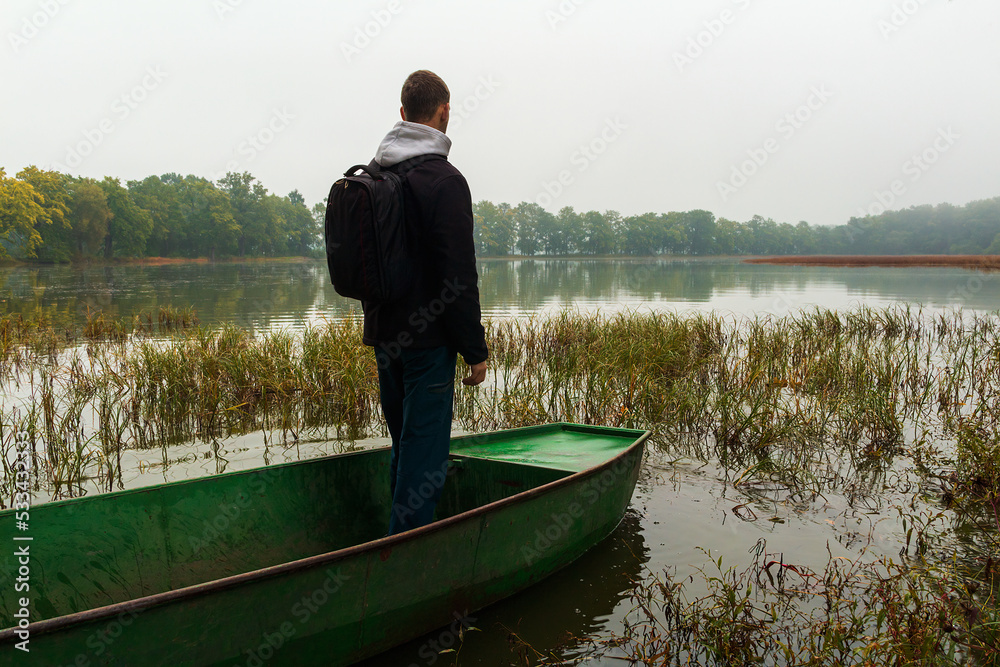Young man in jacket with backpack float on fishing boat at foggy weather. Czech landscape