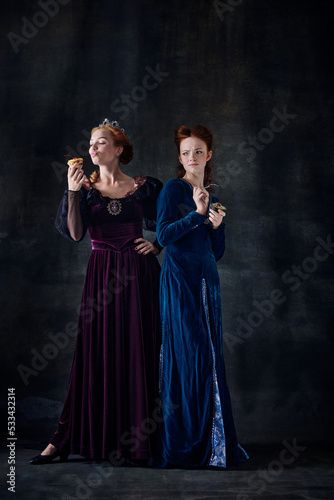 Portrait of two beautiful women, royal persons eating cake and avocado isolated over dark background. Junk and healthy food
