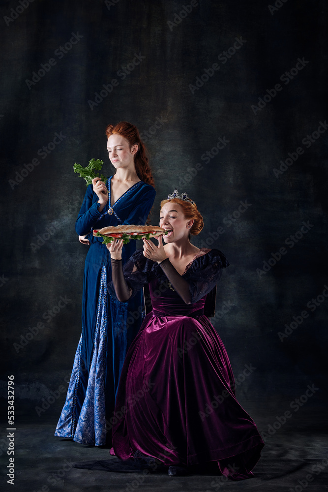 Portrait of two beautiful women, queen eating sandwich, princess eating lettuce isolated over dark background Portrait of two beautiful women, queen eating sandwich, princess eating lettuce isolated over dark background