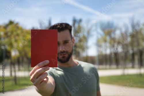 Young and handsome man, with a beard and green shirt, showing a red card with a serious gesture. Concept sport, expulsion, rejection, football, world, competition. Selective focus.