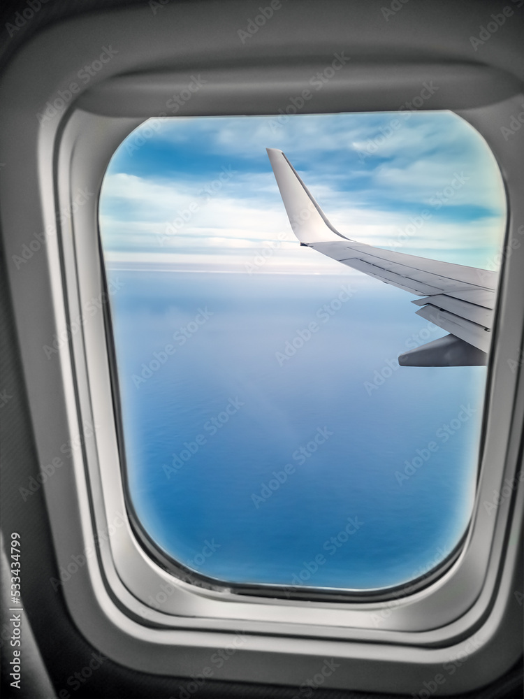 View from the window of the plane on the landscape outside. Oval window frame and airplane wing outside on blue background View from the window of the plane on the landscape outside. Oval window frame and airplane wing outside on blue background