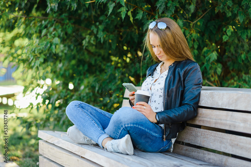 A smiling modern young woman sitting on bench in green park on summer day and talking on smartphone with coffee cup.Girl sitting with a phone and coffe at park.