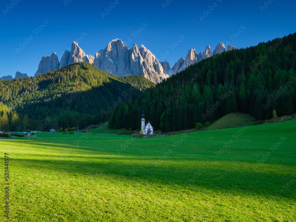 Santa Maddalena. Val di Funes. Dolomite Alps. Italy. Church in the meadow. The mountains and the forest before sunset. Natural landscape in the summertime. Photo in high resolution.
