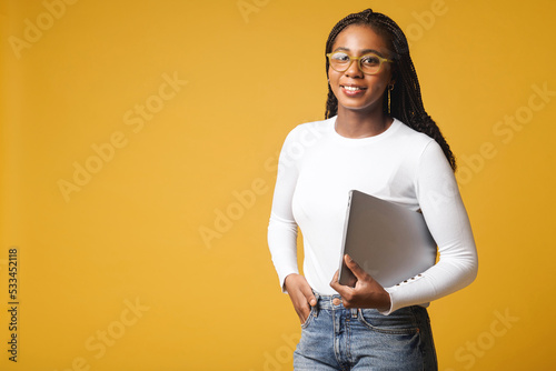 Young African American woman wearing stylish eyeglasses standing isolated on yellow, carrying laptop computer, smiling, female office employee, student photo