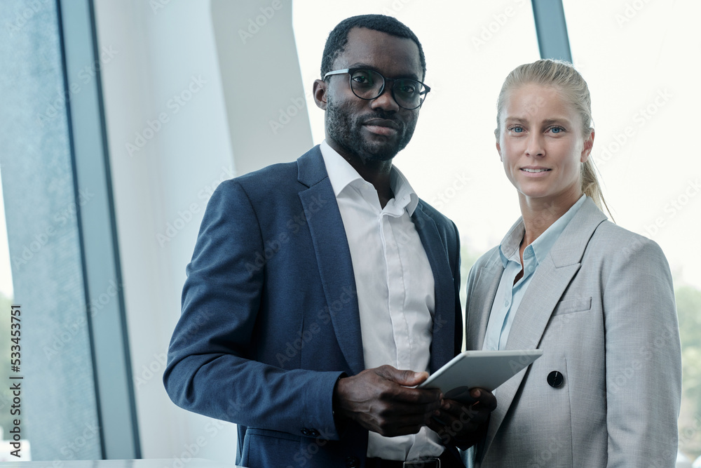 Portrait of two young intercultural office managers or business partners in elegant suits looking at camera while preparing presentation Portrait of two young intercultural office managers or business partners in elegant suits looking at camera while preparing presentation