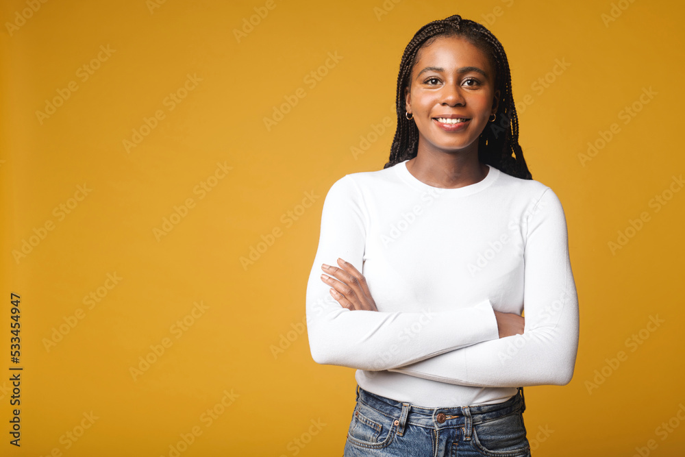Well-looking lady with brunette curly hair looking at camera and smiles, holding arms crossed. Waist up portrait view of the charming smiling woman isolated on yellow background Well-looking lady with brunette curly hair looking at camera and smiles, holding arms crossed. Waist up portrait view of the charming smiling woman isolated on yellow background