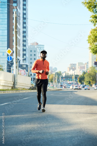Front view of young African American athlete in red sport jacket, black leggiins, sneakers and baseball cap running along asphalt road © pressmaster