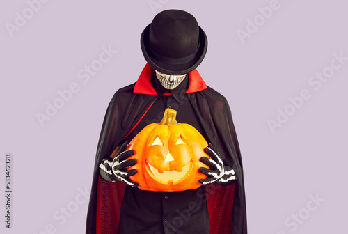 Studio shot of man in skeleton costume holding jack-o-lantern. Man wearing black hat, black and red cloak and gloves standing isolated on light purple background and holding orange Halloween pumpkin