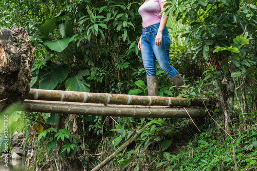 latin peasant woman in marsh boots crossing a bridge made of giant bamboo to cross a stream  surrounded by nature. girl on her way home exploring and hiking. outdoor hike