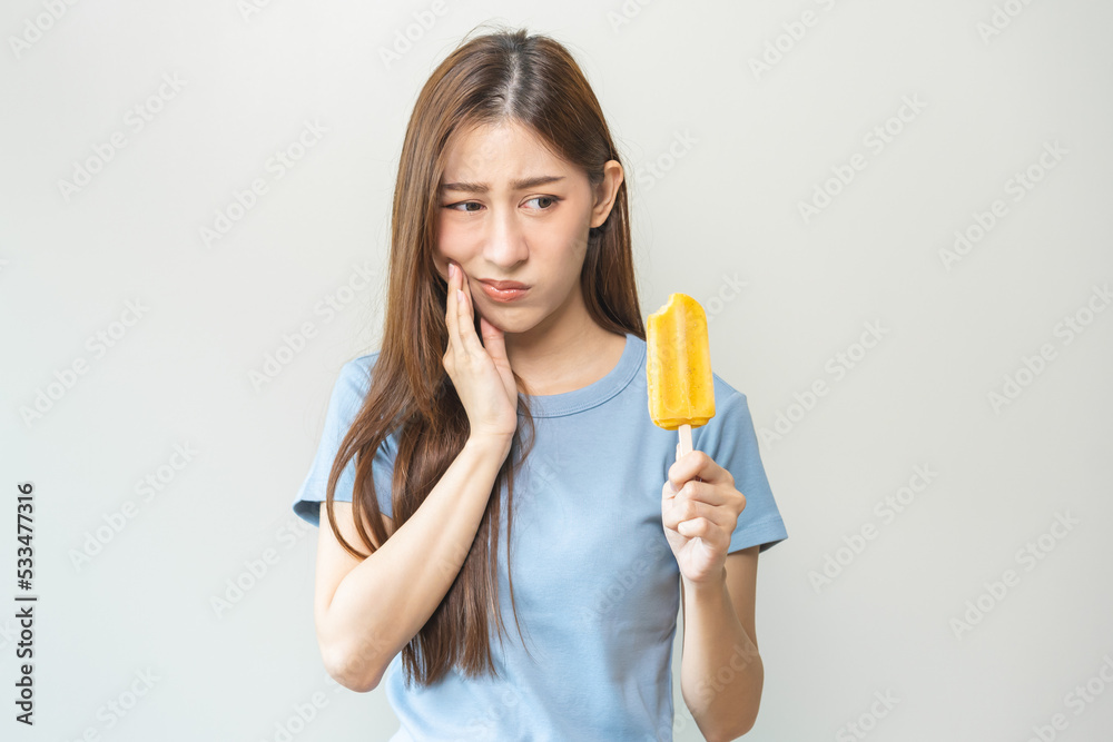 Face expression suffering from sensitive teeth and cold, asian young woman, girl hand touching her cheek, feeling hurt, pain eating ice cream, lolly. Toothache molar tooth at home, dental problem. Face expression suffering from sensitive teeth and cold, asian young woman, girl hand touching her cheek, feeling hurt, pain eating ice cream, lolly. Toothache molar tooth at home, dental problem.