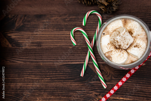 Hot chocolate with marshmallow and coco powder, candy cane pine cones, ribbon, pine needles, pine branch, dark wood, Christmas concept, top view, copy space.