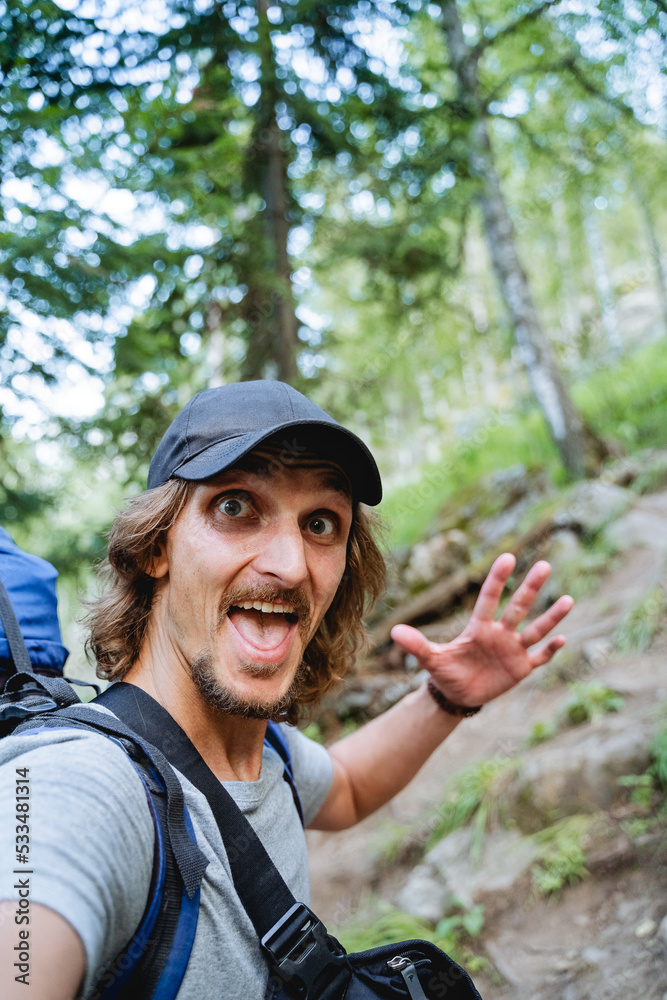 A selfie of a guy on a hike with a backpack on a mountain road, a man smiling at the camera, a large portrait of a hipster in a cap, a photo of himself on his phone, delight, joy