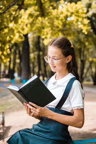 Schoolgirl with glasses reading a book on a warm day in the park. Vertical view