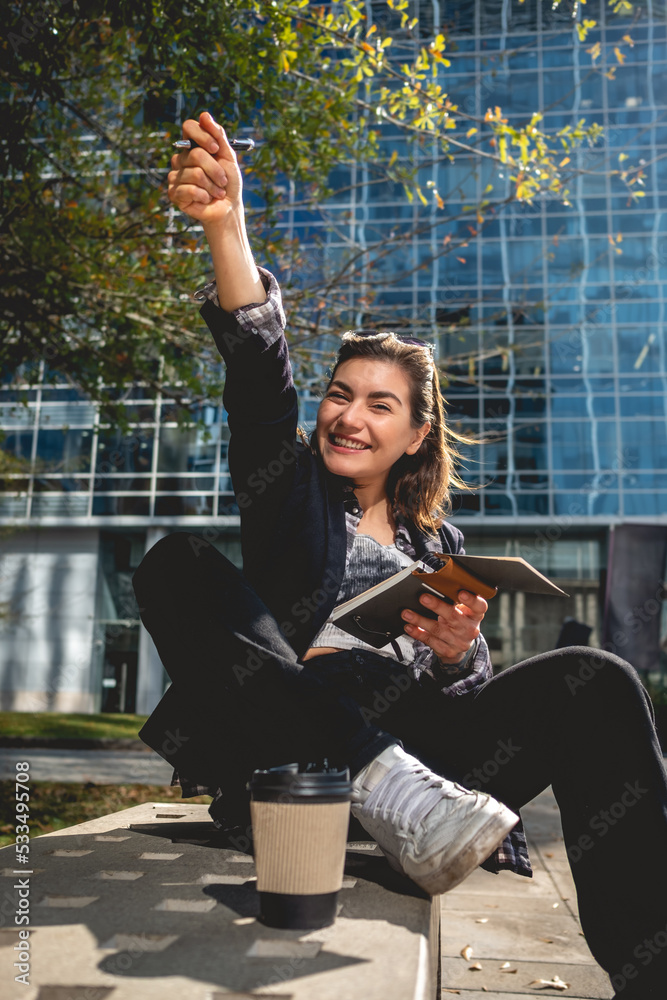 Fototapeta premium Young, successful and happy latina businesswoman with sunglasses and a cup of coffee taking notes of their ideas in a notebook in a park between skyscrapers