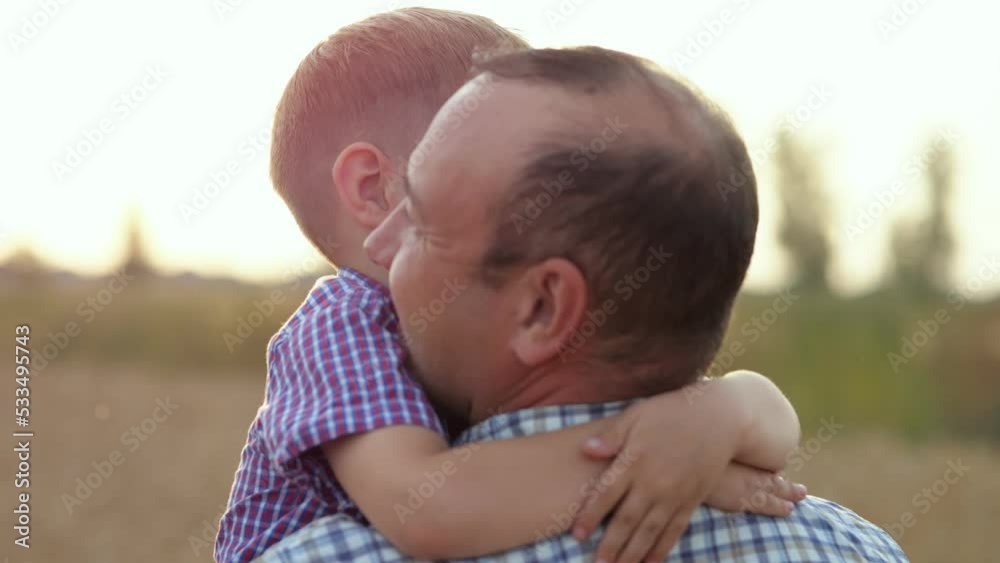Preschooler boy hugging happy father enjoys spending summer weekend in countryside. Smiling son walks in parent arms on wheat field at sunset