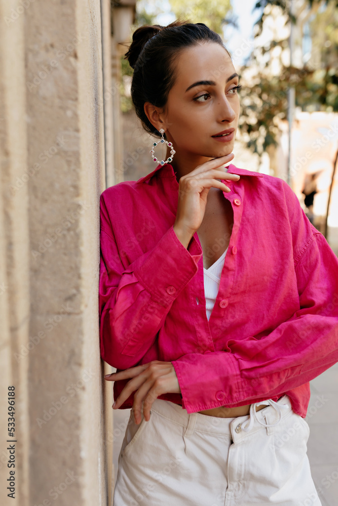 Stylish young caucasian girl looking aside spending free time outdoors. In summer, brunette wears pink blouse and white shorts. Lifestyle concept