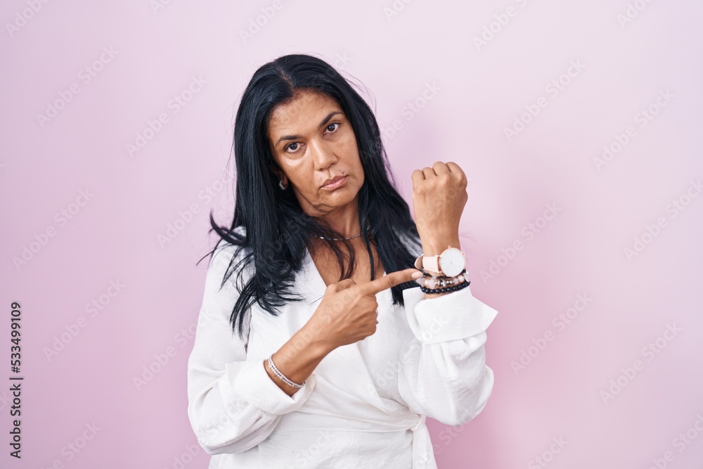 Mature hispanic woman standing over pink background in hurry pointing to watch time, impatience, looking at the camera with relaxed expression Mature hispanic woman standing over pink background in hurry pointing to watch time, impatience, looking at the camera with relaxed expression