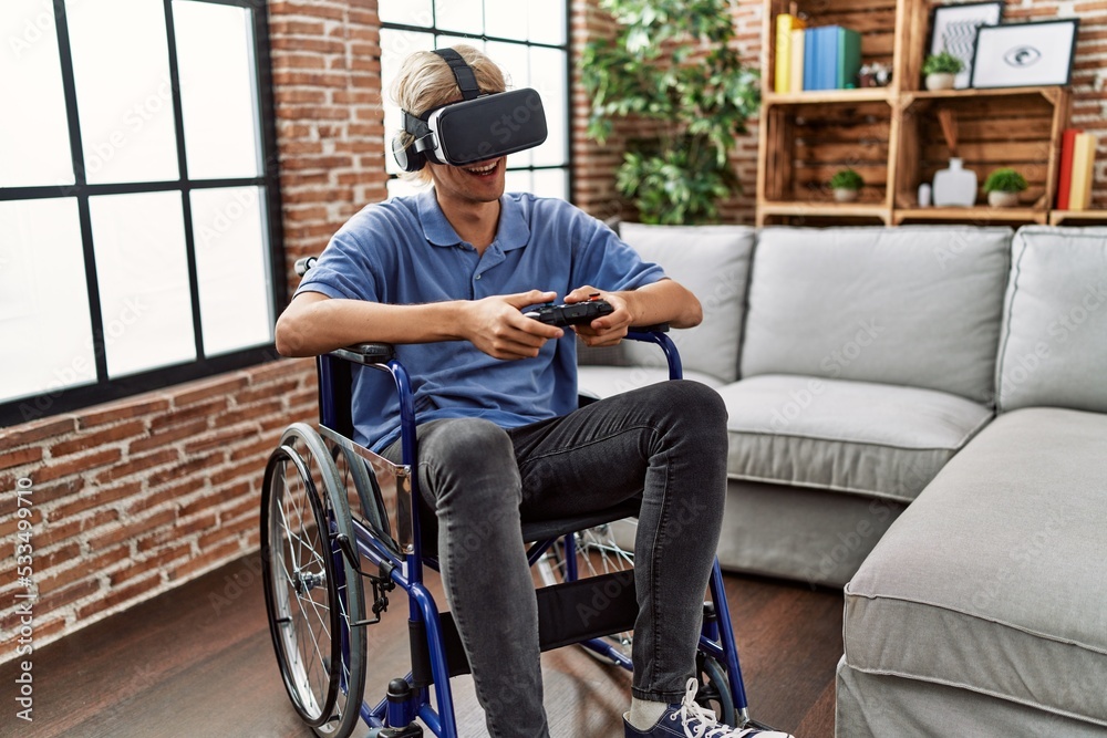 Young blond man playing video game using virtual reality glasses and joystick sitting on wheelchair at home