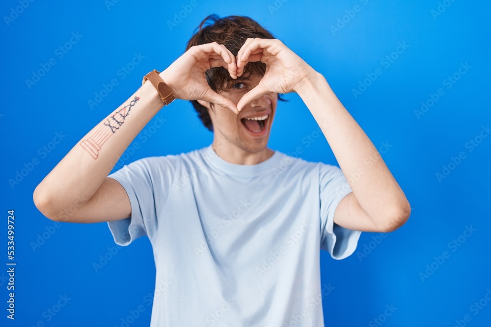 Hispanic young man standing over blue background doing heart shape with hand and fingers smiling looking through sign Hispanic young man standing over blue background doing heart shape with hand and fingers smiling looking through sign