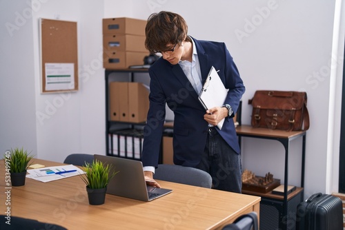 Young blond man business worker holding clipboard using laptop at office