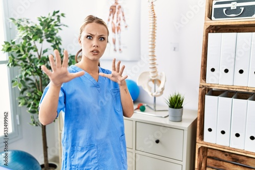 Young caucasian woman working at pain recovery clinic afraid and terrified with fear expression stop gesture with hands  shouting in shock. panic concept.