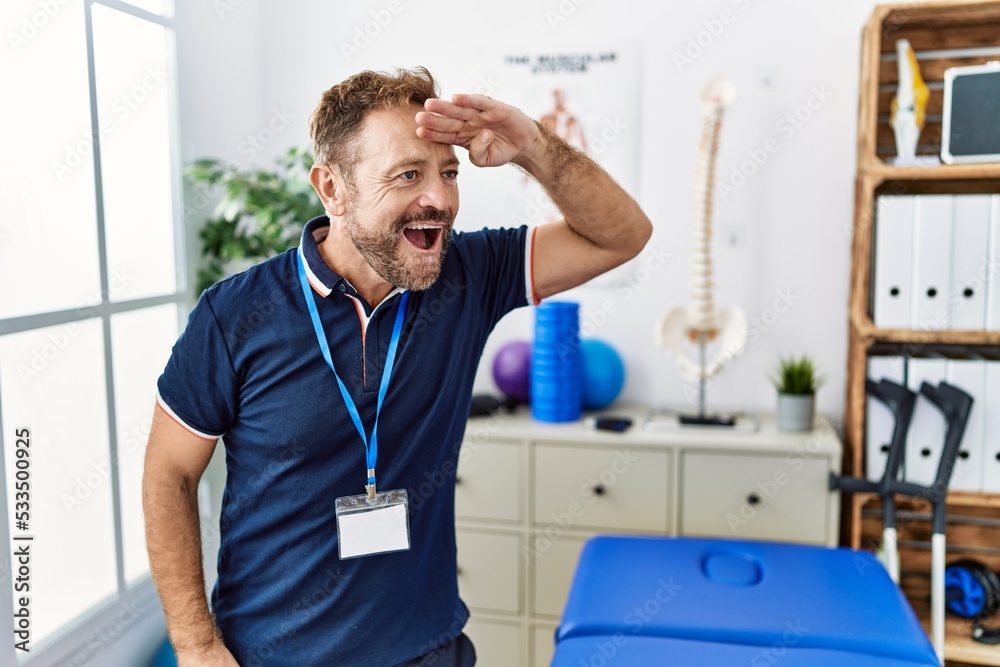 Middle age physiotherapist man working at pain recovery clinic very happy and smiling looking far away with hand over head. searching concept.