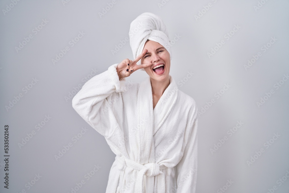 Blonde caucasian woman wearing bathrobe doing peace symbol with fingers over face, smiling cheerful showing victory Blonde caucasian woman wearing bathrobe doing peace symbol with fingers over face, smiling cheerful showing victory
