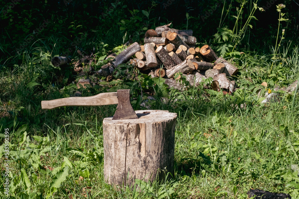 pile of sawn firewood on a background of green grass and trees on a sunny summer day with darkening shadows and a log with an ax in the foreground. Deforestation and ecology concept pile of sawn firewood on a background of green grass and trees on a sunny summer day with darkening shadows and a log with an ax in the foreground. Deforestation and ecology concept