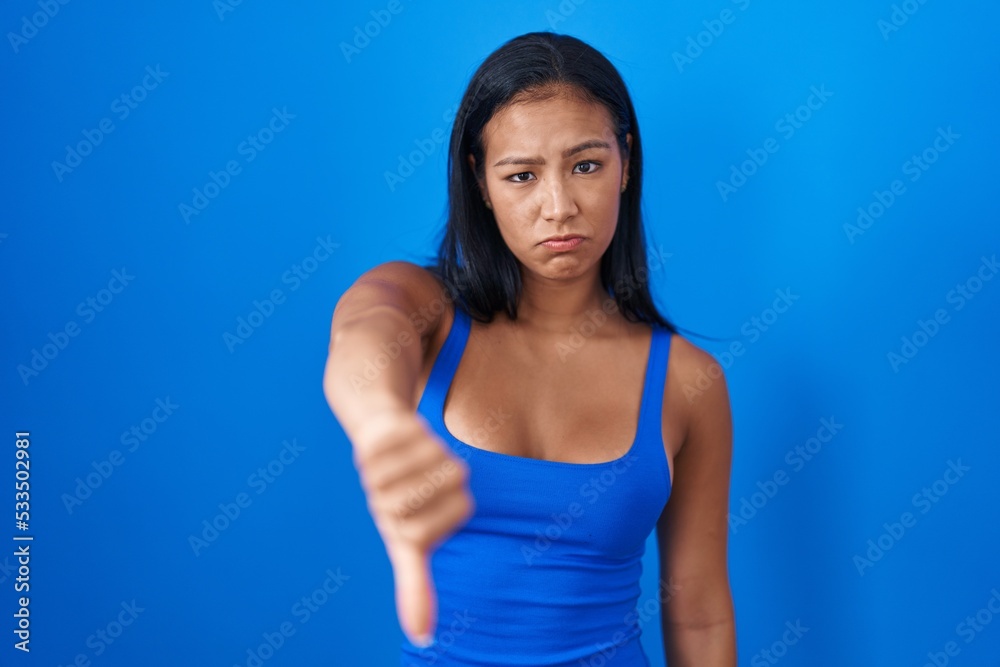 Hispanic woman standing over blue background looking unhappy and angry showing rejection and negative with thumbs down gesture. bad expression. Hispanic woman standing over blue background looking unhappy and angry showing rejection and negative with thumbs down gesture. bad expression.