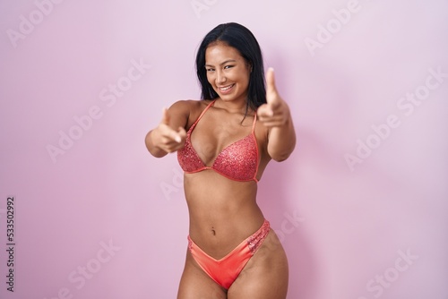 Hispanic woman wearing bikini pointing fingers to camera with happy and funny face. good energy and vibes.
