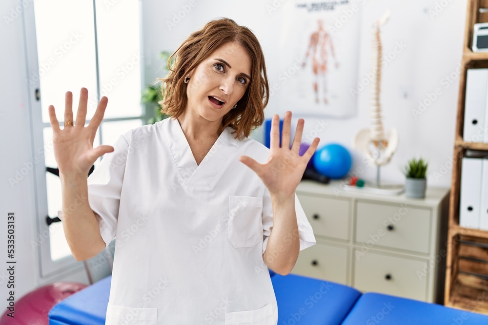Middle age physiotherapist woman working at pain recovery clinic afraid and terrified with fear expression stop gesture with hands, shouting in shock. panic concept. Middle age physiotherapist woman working at pain recovery clinic afraid and terrified with fear expression stop gesture with hands, shouting in shock. panic concept.