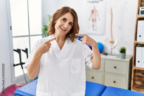 Middle age physiotherapist woman working at pain recovery clinic smiling cheerful showing and pointing with fingers teeth and mouth. dental health concept.