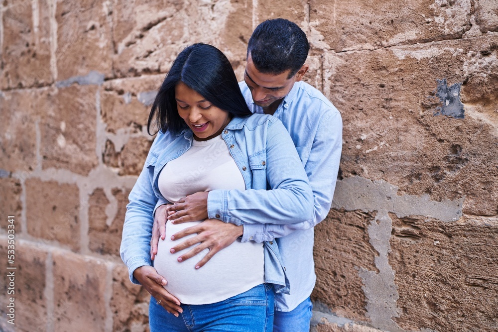 Young latin couple expecting baby touching belly at street