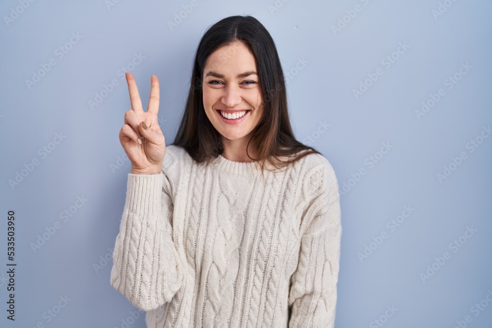 Young brunette woman standing over blue background showing and pointing up with fingers number two while smiling confident and happy. Young brunette woman standing over blue background showing and pointing up with fingers number two while smiling confident and happy.